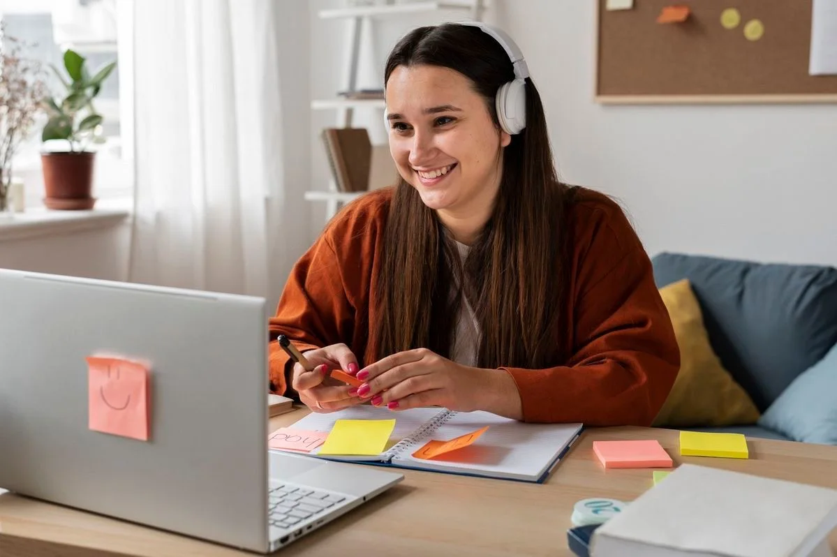 A smiling female student wearing headphones working on a laptop at a bright, organized desk with colorful sticky notes and a notebook